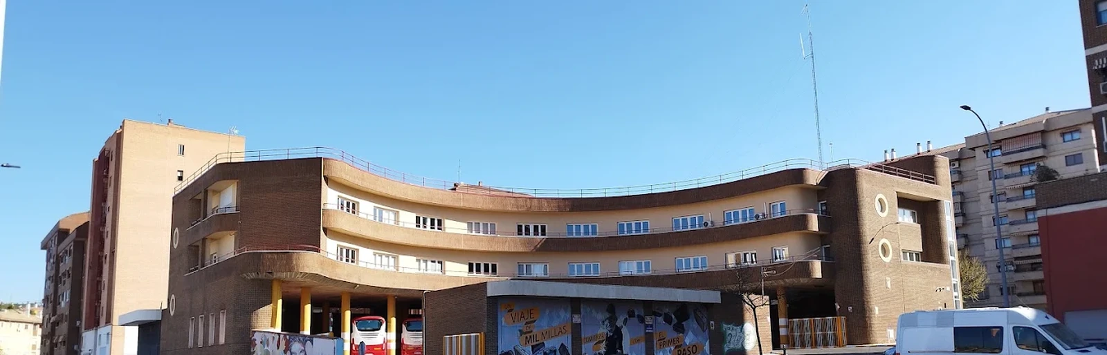 Estación de autobuses de Cuenca vista desde atrás: el edificio curvo de ladrillo con las dársenas cubiertas en la planta baja y dos autobuses estacionados